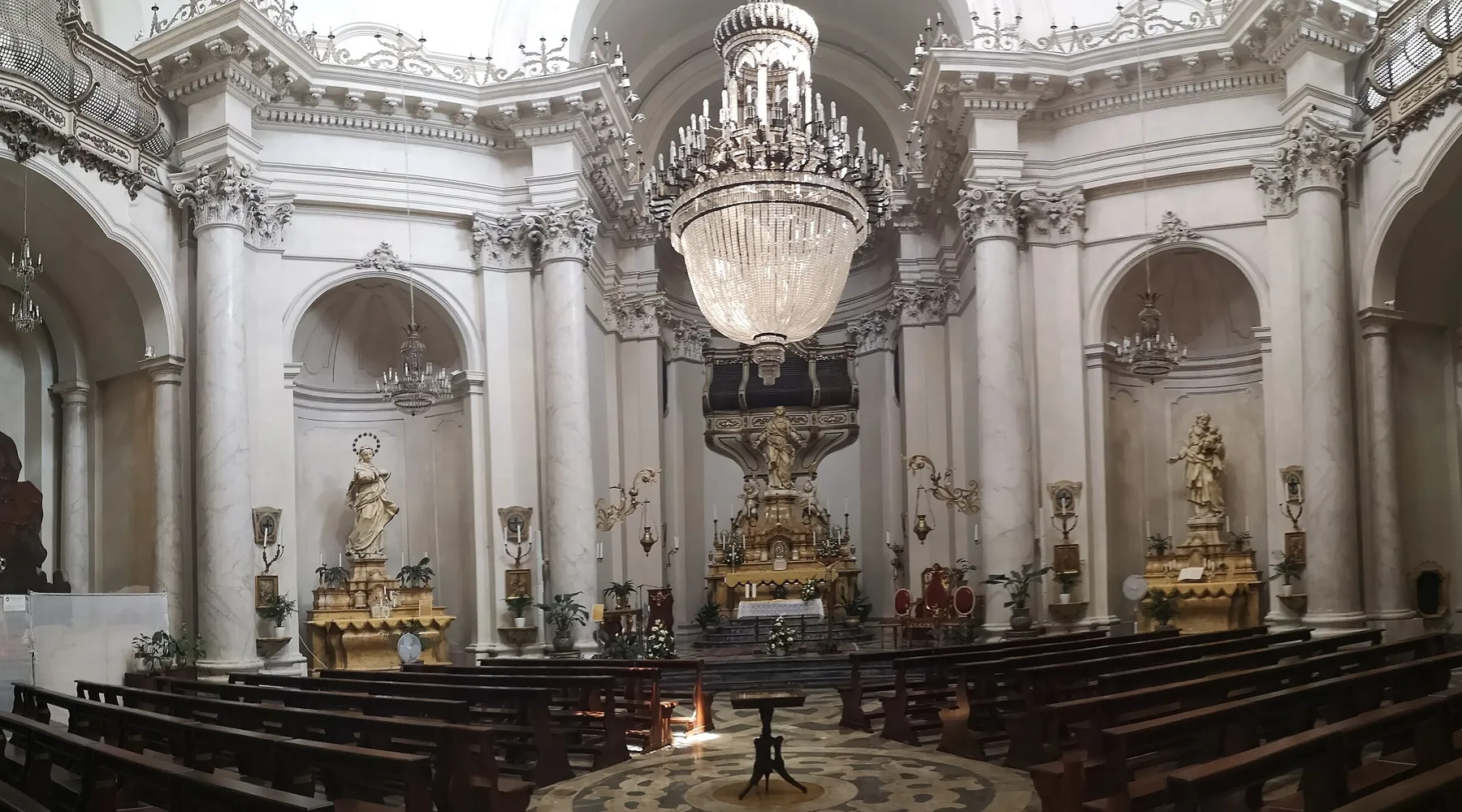 The nave of the Badia di Sant'Agata, with white marble columns, a crystal chandelier, wooden pews and a gilded altar.