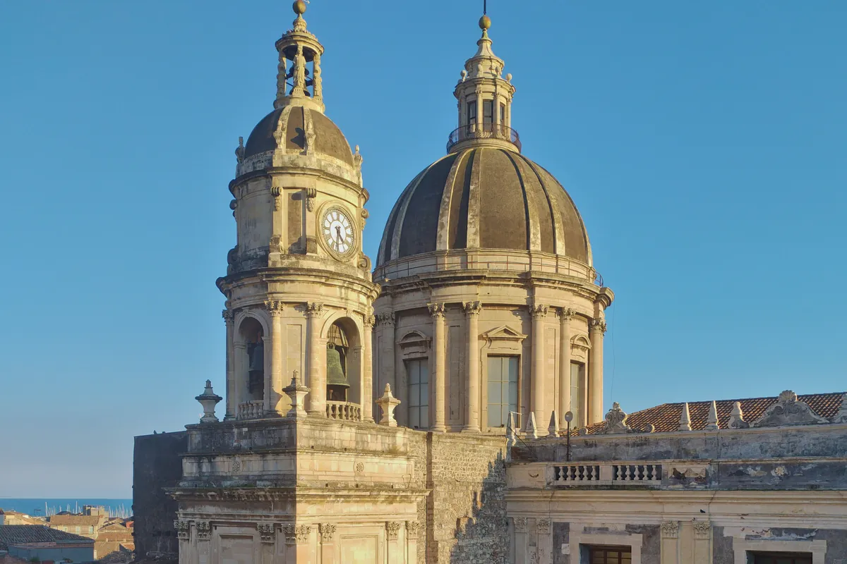 The two domes of Catania Cathedral and its bell tower with a clock, seen from a high terrace against a blue sky